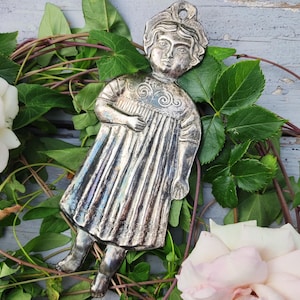 Könnte beinhalten: Ein metallischer, silberfarbener Wandbehang in Form einer stilisierten Figur, die ein plissiertes Kleid mit Spiralmustern am Ausschnitt trägt. Die Figur hat einen runden Kopf mit ausgeprägten Gesichtszügen und ein Loch zum Aufhängen.