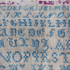 Op de afbeelding: Een sampler met het alfabet geborduurd in blauwe draad op een witte linnen achtergrond.