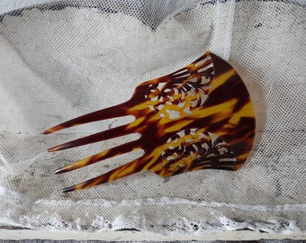 May include: A vintage hair comb with a tortoiseshell pattern. The comb features a curved top with decorative cutouts and three long, pointed tines. The color is a mix of brown and yellow, with a glossy finish. The comb is likely made of celluloid or a similar material.