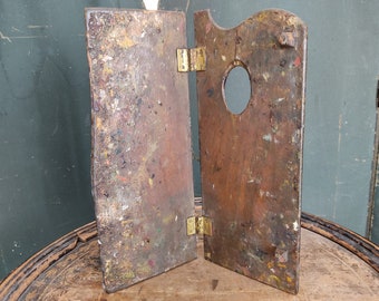 May include: A vintage wooden artist's palette with two hinged panels. The palette is covered in dried paint splatters and has a circular hole in the center.