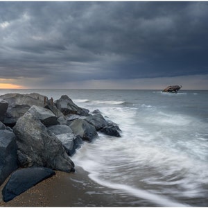 May include: A dramatic seascape featuring a dark, cloudy sky over a rocky shoreline. Large, dark gray boulders line the beach, with the ocean waves creating a blurred, white effect. A shipwreck is visible in the distance. The image is signed by Jason Gambone.