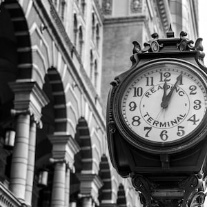 May include: Black and white image of a vintage clock with the words "Reading Terminal" on the face. The clock is mounted on a decorative post. The background features a building with arched windows and columns.