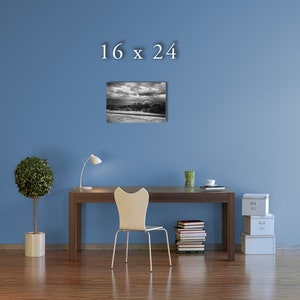 May include: A black and white photograph of a cityscape with a cloudy sky. The image is framed and hanging on a blue wall above a desk with a chair, a lamp, and a stack of books.