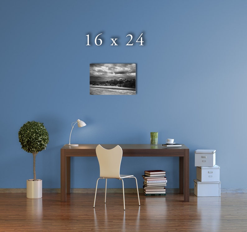 May include: A black and white photograph of a cityscape with a cloudy sky. The image is framed and hanging on a blue wall above a desk with a chair, a lamp, and a stack of books.