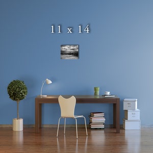 May include: A black and white photograph of a beach scene with a cloudy sky. The image is framed and hanging on a blue wall above a desk with a chair, a lamp, and a stack of books. The text "11 x 14" is printed above the image.