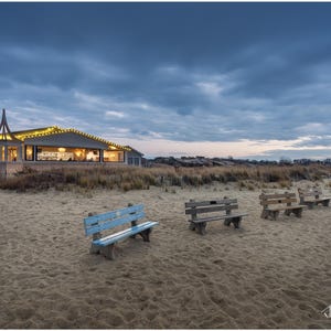 May include: A view of a beach with a building in the background and several wooden benches facing the ocean. The sky is a light blue with clouds. The sand is light brown and the benches are weathered.
