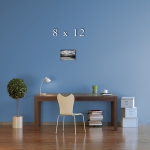 May include: A black and white photo of a landscape with a cloudy sky. The photo is framed and hanging on a blue wall above a desk with a chair, a lamp, and a stack of books. The text "8 x 12" is written in white above the photo.