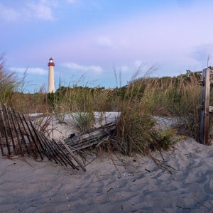 May include: A white lighthouse with a red roof stands tall against a blue sky with clouds. The lighthouse is surrounded by tall grass and a wooden fence. The scene is captured at sunset, with the sky a soft purple and pink.