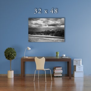 May include: A black and white photograph of a cityscape with a stormy sky. The image is framed and hanging on a blue wall above a desk with a chair, a lamp, and a stack of books.