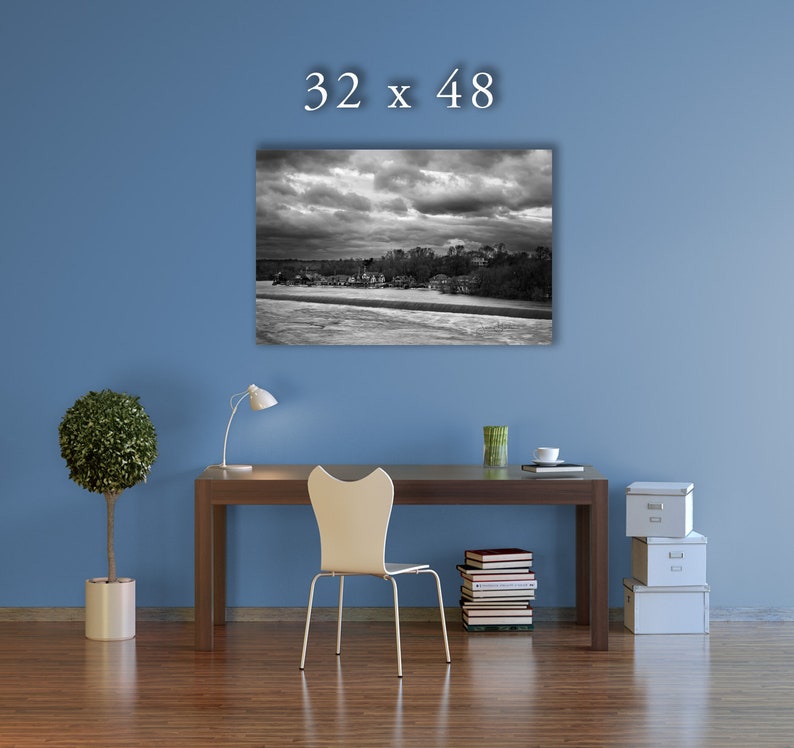 May include: A black and white photograph of a cityscape with a stormy sky. The image is framed and hanging on a blue wall above a desk with a chair, a lamp, and a stack of books.