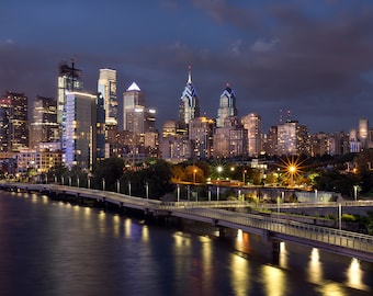 May include: Nighttime cityscape of Philadelphia, Pennsylvania, with illuminated skyscrapers and buildings. The image shows the city skyline reflecting in the dark water, with a bridge and lights in the foreground. The sky is a deep blue.