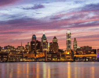May include: A panoramic view of the Philadelphia skyline at dusk, with the city's skyscrapers illuminated against a vibrant pink and purple sky. The reflection of the city lights can be seen in the calm water of the Delaware River.