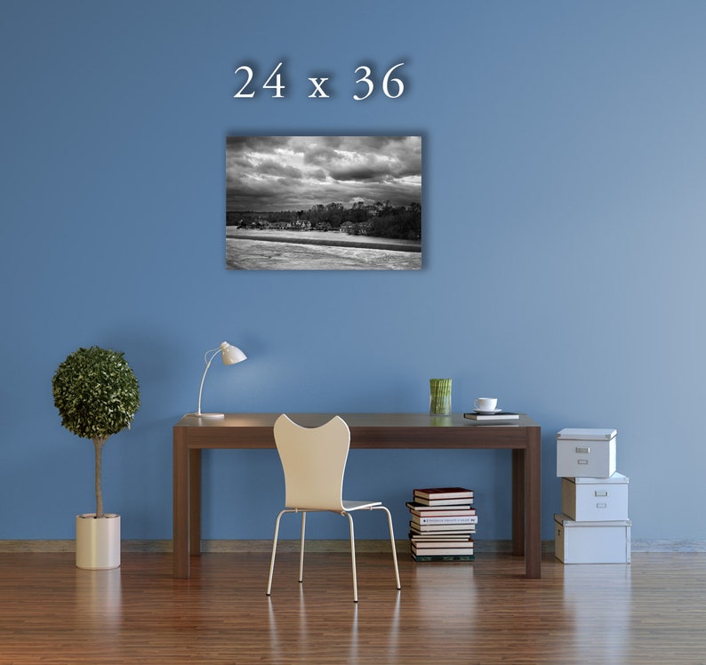 May include: A black and white photograph of a cityscape with a stormy sky. The photo is framed and hanging on a blue wall above a desk with a chair, a lamp, and a potted plant. The text "24 x 36" is printed above the photo.