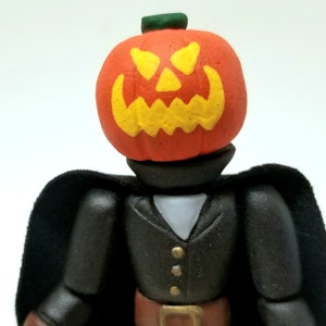 Puede incluir: Una figura de Halloween negra y naranja con cabeza de calabaza y una capa negra. La cabeza de calabaza tiene una cara tallada amarilla.