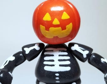 Puede incluir: Una figura de esqueleto blanco y negro con una cabeza de calabaza. La calabaza es naranja con una cara sonriente amarilla.