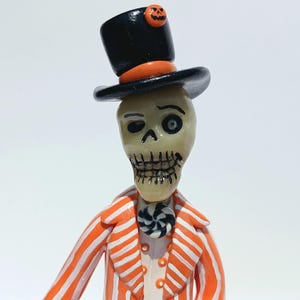 May include: A figurine of a skeleton wearing a black top hat with an orange band and a small orange jack-o'-lantern accent. The skeleton wears an orange and white striped jacket with a black and white spiral button at the neck and small buttons.