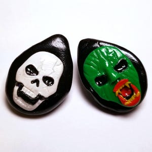 May include: Two Halloween-themed earrings, one with a white skull on a black background and the other with a green monster face with red and yellow teeth on a black background.