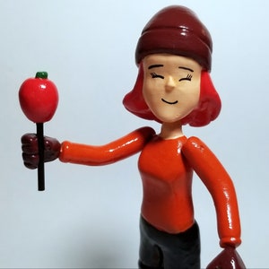 Puede incluir: Una figurita hecha a mano de una persona que lleva un sombrero marrón y un suéter naranja sosteniendo una manzana roja en un palo.