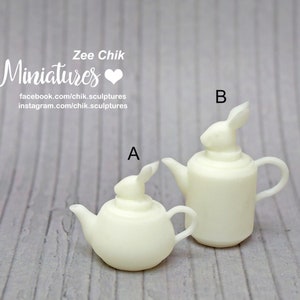 May include: Two white ceramic teapots with rabbit lid finials. The smaller teapot is labelled "A" and the larger teapot is labelled "B".