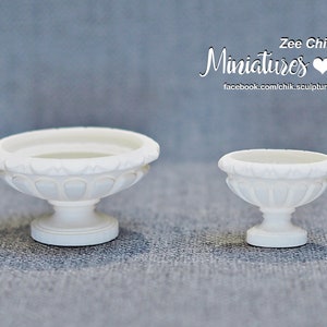 Peut inclure: Deux pots en céramique blanche, l'un plus grand que l'autre, avec un motif décoratif autour du bord. Les deux pots ont une base en piédestal.