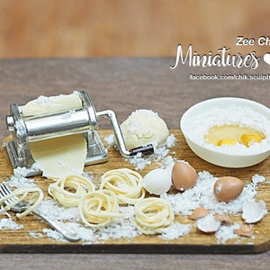 May include: A miniature pasta maker with a silver roller and a wooden chopping board covered in flour. There are several small bowls of flour, eggs, and a small fork. The text "Zee Chik Miniatures" is visible in the top right corner of the image.