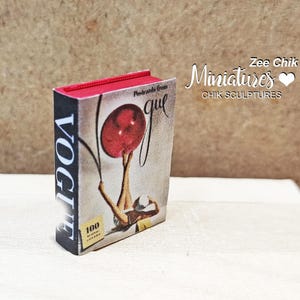 Pode incluir: Uma r&eacute;plica em miniatura da capa da revista Vogue que mostra uma mulher segurando uma bola vermelha. A capa diz "Vogue" e "100 anos de glamour".