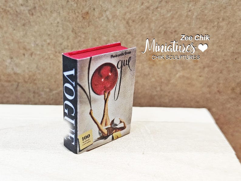 Pode incluir: Uma r&eacute;plica em miniatura da capa da revista Vogue que mostra uma mulher segurando uma bola vermelha. A capa diz "Vogue" e "100 anos de glamour".