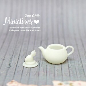Pu&ograve; includere: Una teiera in miniatura di ceramica bianca e una statuetta di coniglio. La teiera ha un corpo arrotondato e un beccuccio. La statuetta di coniglio &egrave; in piedi su una piccola base. Il testo "Zee Chik Miniatures" &egrave; visibile nell'immagine.