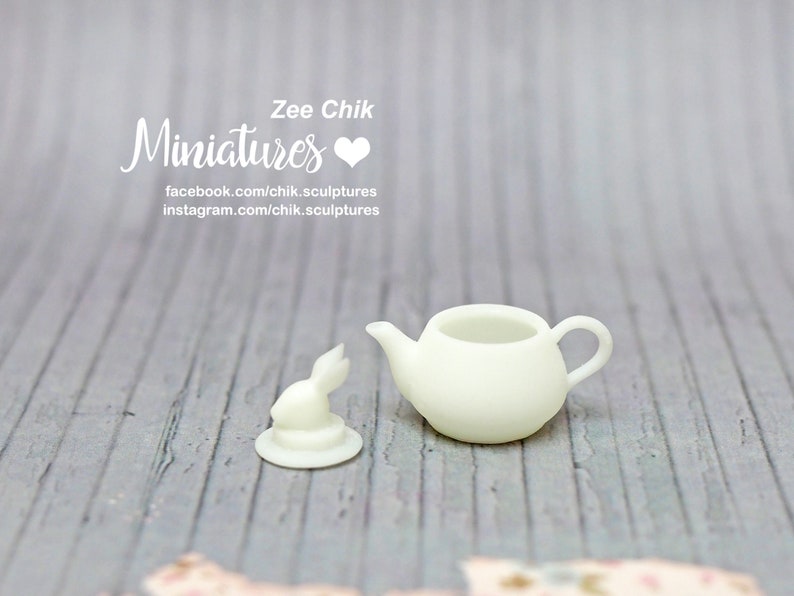 Pu&ograve; includere: Una teiera in miniatura di ceramica bianca e una statuetta di coniglio. La teiera ha un corpo arrotondato e un beccuccio. La statuetta di coniglio &egrave; in piedi su una piccola base. Il testo "Zee Chik Miniatures" &egrave; visibile nell'immagine.