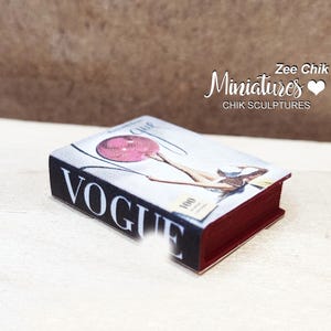 Pode incluir: Uma r&eacute;plica em miniatura da revista Vogue com uma lombada vermelha e uma capa com uma mulher segurando um bal&atilde;o rosa. O texto da capa diz "Vogue" e "1000".