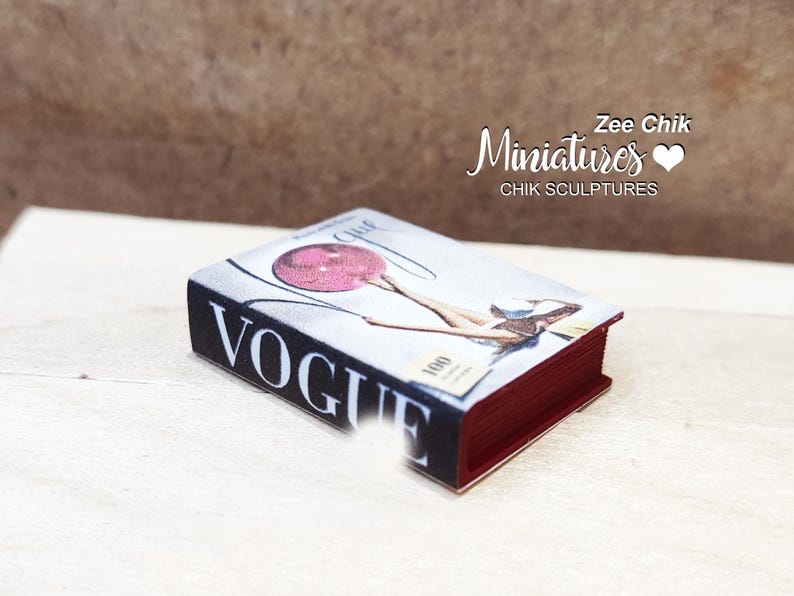 Pode incluir: Uma r&eacute;plica em miniatura da revista Vogue com uma lombada vermelha e uma capa com uma mulher segurando um bal&atilde;o rosa. O texto da capa diz "Vogue" e "1000".