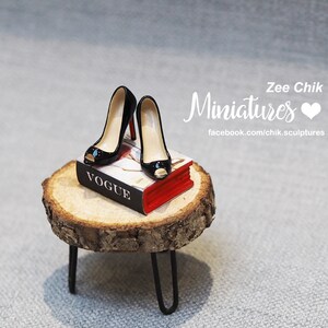 Pode incluir: Um par de sapatos pretos de salto alto em miniatura com dedos a vista s&atilde;o colocados em cima de um livro vermelho e branco intitulado "Vogue". O livro est&aacute; sentado em um pequeno toco de madeira com um suporte de metal preto.