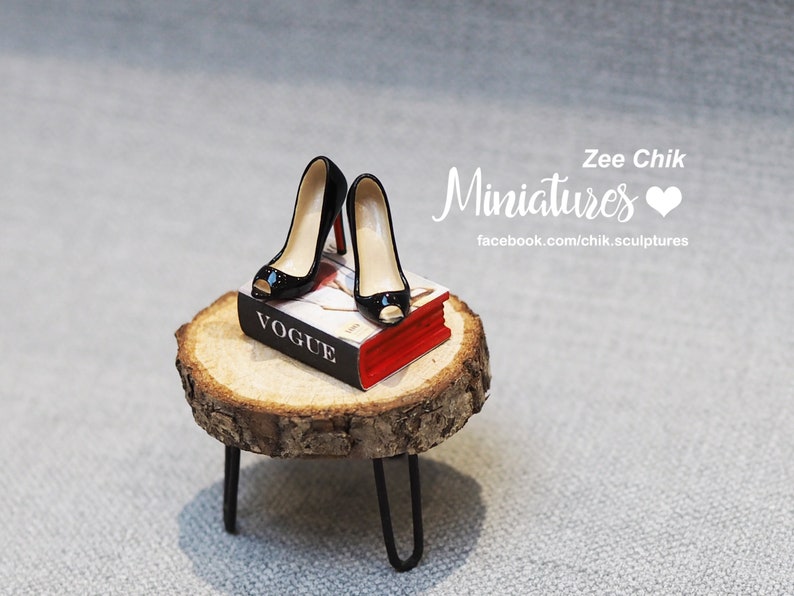 Pode incluir: Um par de sapatos pretos de salto alto em miniatura com dedos a vista s&atilde;o colocados em cima de um livro vermelho e branco intitulado "Vogue". O livro est&aacute; sentado em um pequeno toco de madeira com um suporte de metal preto.