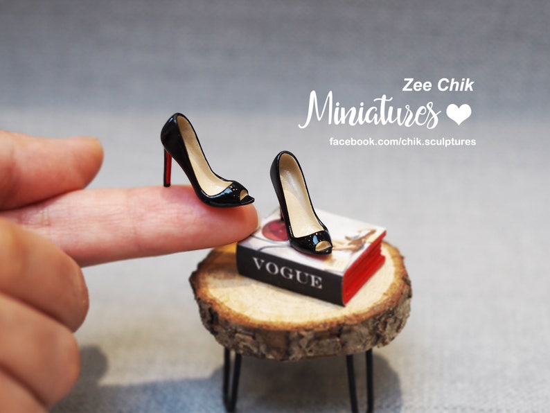 Pode incluir: Dois sapatos pretos de salto alto em miniatura com solas vermelhas. Um sapato est&aacute; em um dedo, o outro est&aacute; em um livro em miniatura intitulado "Vogue".