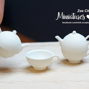 May include: White ceramic miniature teapot and teacup set. The teapot has a round body and a lid with a knob. The teacup has a rounded shape and a handle. The set is on a wooden surface.