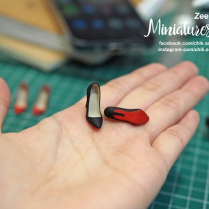 May include: A hand holds two miniature black high-heeled shoes with red soles. The shoes are very small, about the size of a fingernail. The background is blurred, but you can see a green mat and other miniature shoes.