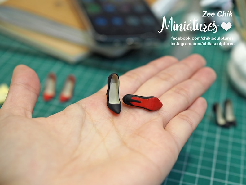 May include: A hand holds two miniature black high-heeled shoes with red soles. The shoes are very small, about the size of a fingernail. The background is blurred, but you can see a green mat and other miniature shoes.