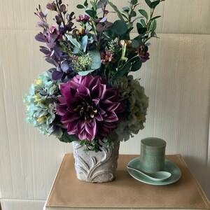 Puede incluir: Un ramo de flores moradas y azules en un jarrón de cerámica blanca con un diseño ornamentado. El jarrón está sobre una superficie marrón claro.