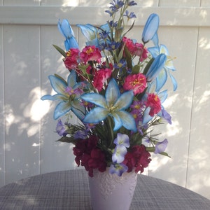 May include: A bouquet of artificial flowers in a lavender vase. The bouquet includes blue, pink, and purple flowers. The vase has a decorative design.