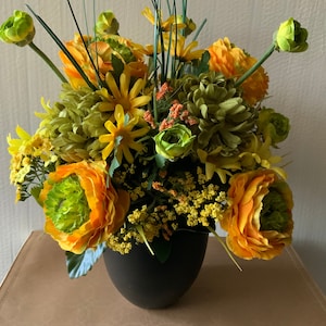 May include: A black vase filled with a floral arrangement of yellow and green flowers. The flowers are arranged in a circular pattern and include yellow roses, green carnations, and yellow daisies.
