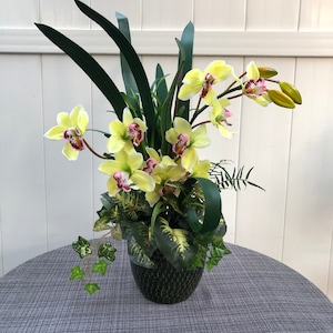 May include: A yellow and pink orchid flower arrangement in a green ceramic vase. The arrangement is surrounded by green leaves and is sitting on a gray table.