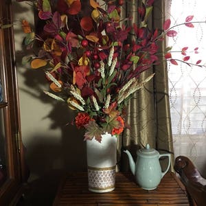 May include: A white vase with a brown and gold pattern holds a bouquet of fall foliage with red berries, orange flowers, and green and brown leaves. A teal teapot sits on a wooden table next to the vase.