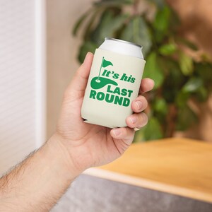 May include: A white can cooler with a green golf course and flag graphic. The text "It's his last round" is printed on the cooler.
