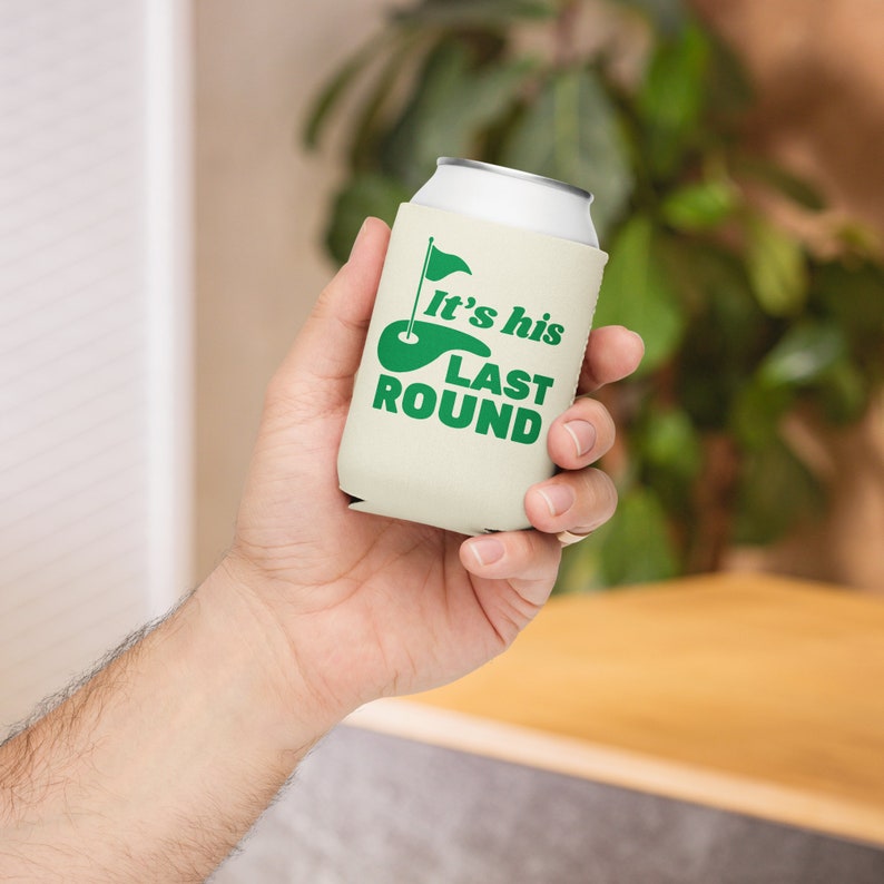 May include: A white can cooler with a green golf course and flag graphic. The text "It's his last round" is printed on the cooler.
