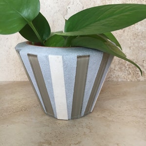 Puede incluir: Un macetero de cerámica rayado en gris y blanco con una planta verde en su interior. El macetero tiene una boca ancha y una base estrecha.