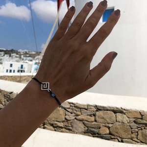 Peut inclure: Un bracelet en cordon noir avec un charme argenté représentant un motif de clé grecque et un œil bleu maléfique. Le bracelet est porté au poignet d'une personne sur fond d'un bâtiment blanc avec un moulin à vent.