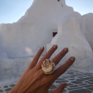 Puede incluir: Un anillo grande y redondo hecho de una concha marina, con una base crema y marcas de color marrón rojizo. El anillo se lleva en una mano con esmalte de uñas brillante, sobre un fondo borroso de un edificio blanco y cielo azul.