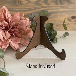 May include: A wooden stand with three legs, designed to hold a plate or other flat object. The stand is brown and has a simple design. The text "Stand Included" is printed below the stand.