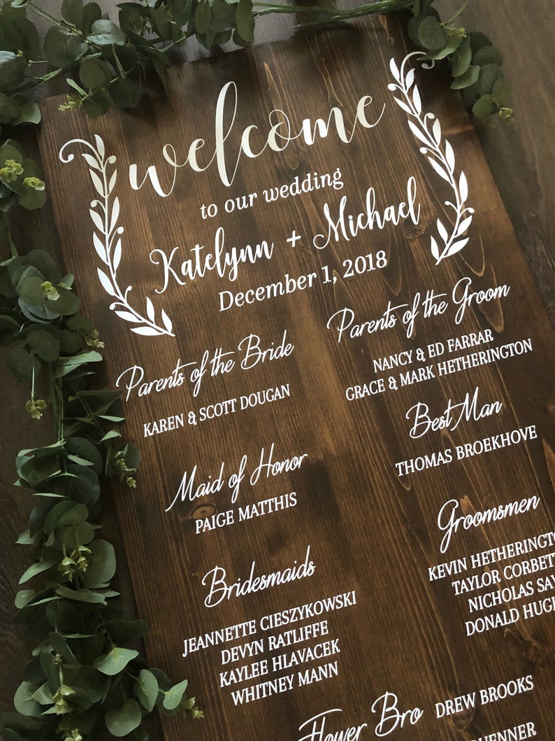 Puede incluir: Un letrero de madera con letras blancas anunciando una boda para Katelynn y Michael el 1 de diciembre de 2018. El letrero enumera los nombres de los padres de la novia y el novio, la dama de honor, las damas de honor, el padrino de boda y los padrinos de boda.