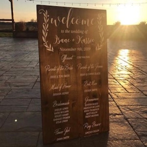 Puede incluir: Un cartel de boda de madera r&uacute;stica con caligraf&iacute;a blanca. El cartel dice "Bienvenido a la boda de Isaac + Kassie, 9 de noviembre de 2019". Tambi&eacute;n enumera al oficiante, los padres de la novia y el novio, la dama de honor, el padrino de honor, las damas de honor, los padrinos de honor, la ni&ntilde;a de las flores y el portador de anillos.